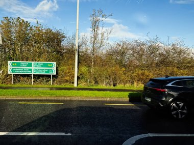 Longford, İrlanda - 21 Ekim 2025: Siyah bir SUV İrlanda 'da ıslak bir yolda, çitleri ve mavi gökyüzü olan yeşil istikamet işaretlerinin yanından geçiyor.
