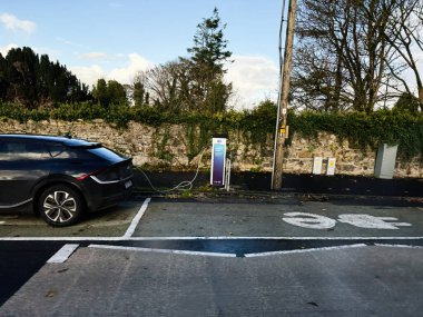 Castlerea, İrlanda - 21 Ekim 2025: İrlanda 'da taş bir duvarın yanındaki yol kenarındaki bir istasyonda siyah bir Kia elektrikli araba şarj oluyor.