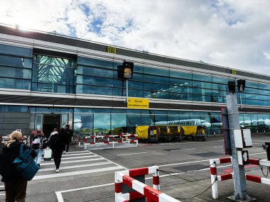 Dublin, İrlanda - 21 Ekim 2025: Yolcular cam cepheli Dublin havaalanı terminaline parlak bir gökyüzünün altında bavul ve çanta taşıyarak yaklaşıyorlar.