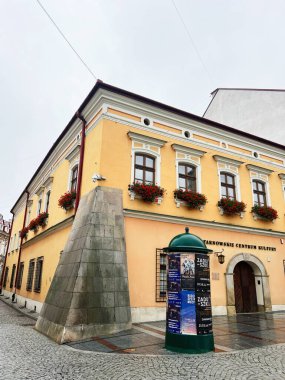 Tarnow, Polonya - 25 Ekim 2025: Çiçek kutuları, taş duvar ve Polonya 'nın Tarnow kentindeki tuhaf kaldırım taşı caddesi boyunca yeşil bir poster kulübesi bulunan tarihi sarı bina. 