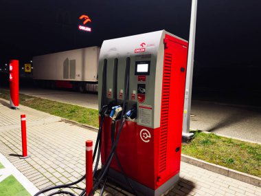 Krakow, Polonya - 07 Kasım 2025: Polonya 'daki gece yakıt ikmali ve şarj istasyonuna kırmızı bir dağıtıcı ve park halindeki bir kamyon römorkunun yanında bağlı kablolar.