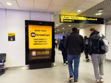 Manchester, İngiltere - 07 Kasım 2025: Manchester havaalanı terminalinde gezginler açık sarı bir tramvay reklamının yanından geçtiler ve.