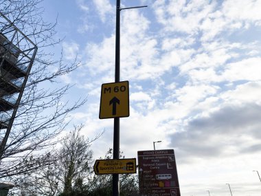 Manchester, İngiltere - 16 Kasım 2025: M60 ok işaretli uzun lamba direği ve mavi gökyüzüne karşı kampüs yön tahtaları.