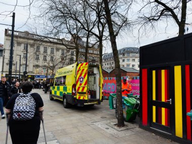 Manchester, İngiltere - 16 Kasım 2025: Manchester caddesindeki sarı ambulans, yayalar, dükkanlar ve manzaralı bir inşaat sahası.