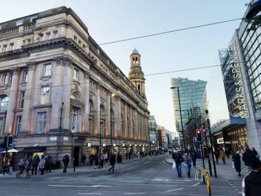 Manchester, İngiltere - 19 Kasım 2025: İnsanlar süslü taş binalar ve cam alışveriş merkezleri olan bir cadde boyunca yürüyüp alışveriş yapıyorlar.