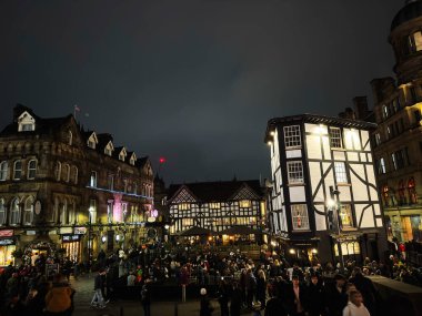 Manchester, İngiltere - 22 Kasım 2025: Kalabalık festival ışıkları ve keresteli binalarla aydınlatılmış tarihi Manchester meydanını gece doldurdu.