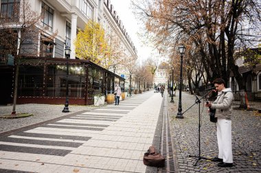 Chisinau, Moldova - 22 Kasım 2025: Bir sokak müzisyeni, sonbaharda ağaçlarla kaplı bir caddede bir kafenin yanında şarkı söylüyor ve gitar çalıyor.