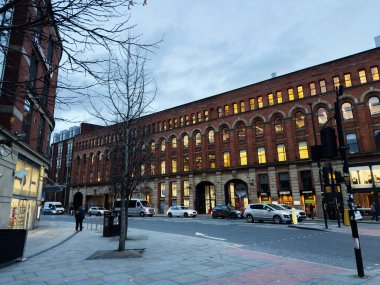 Manchester, İngiltere - 10 Aralık 2025: Manchester, İngiltere 'de şehir manzarası kırmızı tuğla mimari, dükkan önleri ve ışıklı pencereleri yakıyor.