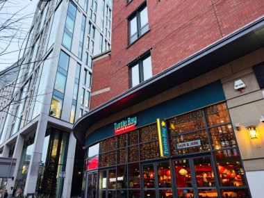 Manchester, İngiltere - 10 Aralık 2025: Sıcak ışıkları ve tuğla binaları olan bir Turtle Bay restoranının yer aldığı canlı bir Manchester sokak sahnesi.
