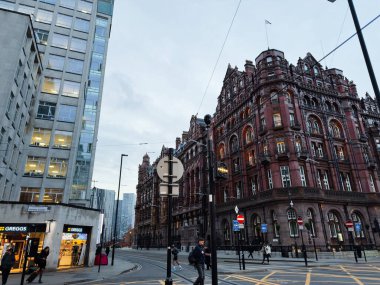 Manchester, İngiltere - 10 Aralık 2025: İnsanlar Manchester, İngiltere 'deki çağdaş ofislerin yanında tarihi tuğla mimarinin yanından geçiyor, tramvay hatları arasında.
