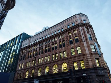 Manchester, İngiltere - 10 Aralık 2025: Manchester, İngiltere 'de tarihi bir tuğla bina, yakınlardaki modern cam yapı yükselirken aydınlatılmış pencereler.