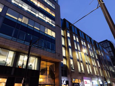 Manchester, İngiltere - 10 Aralık 2025: Aydınlatılmış pencereleri ve tramvay hatları olan kentsel sokak manzaralı çağdaş Manchester binasının akşam manzarası.