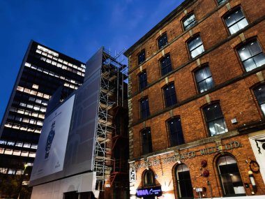 Manchester, İngiltere - 10 Aralık 2025: Manchester 'da gece yarısı şehir caddesi tuğla fabrikası cephe fabrikaları sınırlı, iskele iskelesi ve uzun bir reklam panosu ile