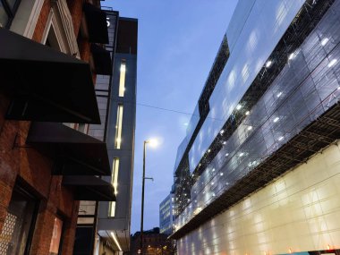 Manchester 'daki alacakaranlık sahnesinde tuğla dükkanları, sokak lambaları ve modern cam inşaatlar gösteriliyor..