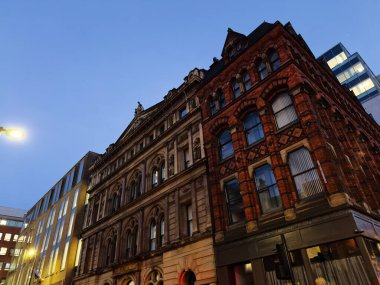 Manchester 'daki şehir sokak sahnesinde mavi saatte modern cam cephe yanında kırmızı tuğlalı tarihi bir bina yer alıyor..