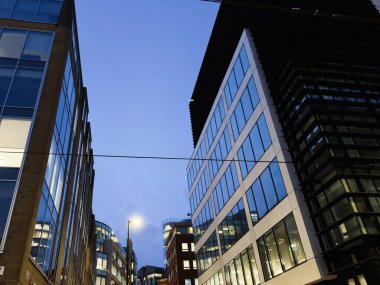 Manchester 'ın çağdaş ofis bloklarının üzerindeki mavi alacakaranlık cam cepheleri ve sokak ışıklarıyla, kentsel bir hava yaratıyor..