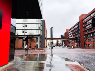 Manchester, İngiltere - Aralık 06, 2025: Yağmurlu bir günde Manchester şehir sokağı; modern kırmızı mağazalar, ıslak kaldırımlar ve şemsiyeli yayalar.