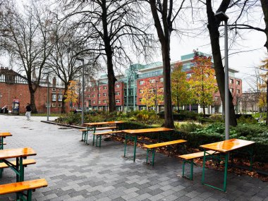 Manchester 'daki sonbahar sahnesinde banklar, ağaçlar, asfalt bir plaza ve çağdaş tuğla kampüs binaları yer alıyor..