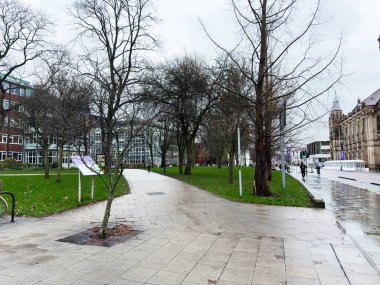 Manchester, İngiltere - Aralık 06, 2025: Yağmurlu bir günde Manchester 'da ıslak asfalt, yapraksız ağaçlar ve yakınlardaki tarihi mimariyle şehir parkı sahnesi.