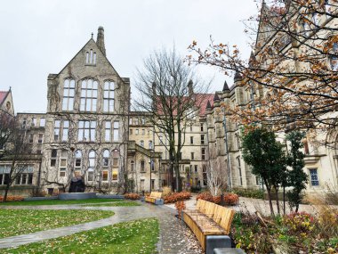 Manchester, İngiltere - Aralık 06, 2025: Sonbahar ışığı altında Gotik taş binası, ağaçları ve bankları olan Manchester Üniversitesi avlusu.