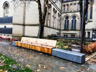 Manchester, UK - December 06, 2025: Wooden bench in a courtyard beside a Manchester church, twinkling lights, autumn leaves, wet pavement.
