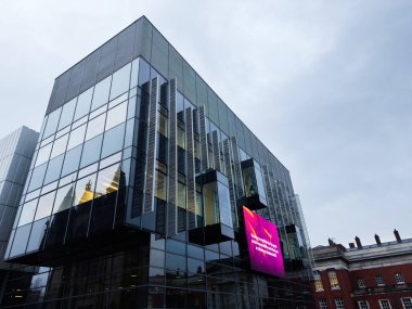 Manchester, İngiltere - Aralık 06, 2025: Manchester UK 'de yansıtıcı pencereler ve gri gökyüzüne karşı cesur dijital tabelalar içeren çağdaş cam cephe.