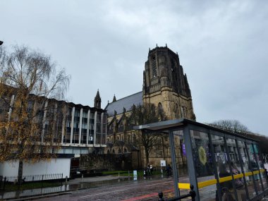 Manchester, İngiltere - Aralık 06, 2025: Manchester 'da yağmurlu bir günde ıslak bir şehir caddesinde modern bir otobüs durağının yanında uzun bir katedral kulesi görülüyor.