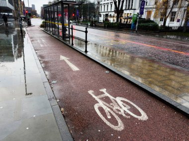 Manchester, İngiltere - 06 Aralık 2025: Manchester 'da yağmurlu bir gün bisiklet yolu, otobüs durağı barınağı ve ıslak kentsel sokaklara yansıyor.