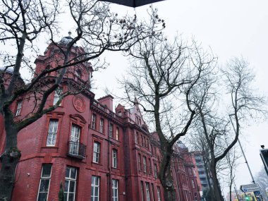 Manchester, İngiltere - Aralık 06, 2025: Manchester 'da gri bir günde yapraksız ağaçlarla çevrili kırmızı tuğlalı bir mimari bina.