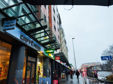 Manchester, İngiltere - Aralık 06, 2025: Manchester 'da Cafe Nero ve Subway tabelalı kentsel mağazalar, yağmurlu bir günde şemsiye altında yayalar.