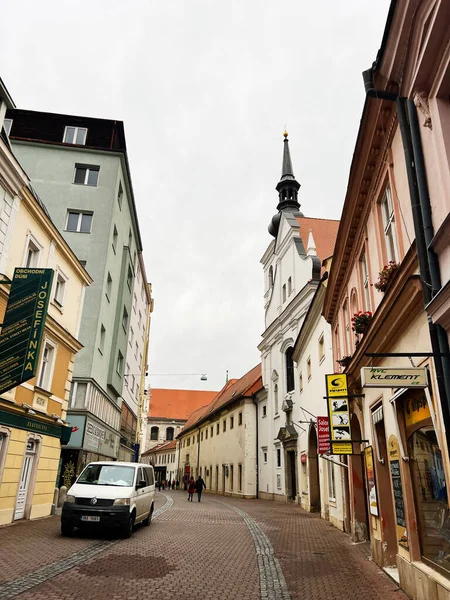 Brno, Çek Cumhuriyeti - 29 Kasım 2025: Tarihi Brno şehrinde sakin bir cadde dükkanları, tabelaları ve kavisli kaldırım taşı yolu üzerinde beyaz bir minibüs.