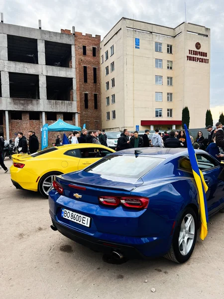 Ternopil, Ukrayna - 29 Kasım 2025: Mavi Chevrolet Camaro ve sarı araba bir üniversite binasının önünde toplanıyor, Ukrayna bayrağı da yakınlarda.