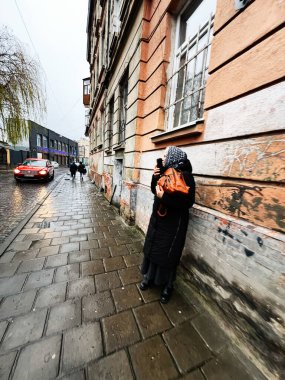 Lviv, Ukrayna - 29 Kasım 2025: Siyah paltolu ve eşarplı yaşlı bir kadın, Ukrayna 'nın Lviv kentindeki yağmurlu bir caddede duvar yazısı duvarında telefonuyla fotoğraf çekiyor..