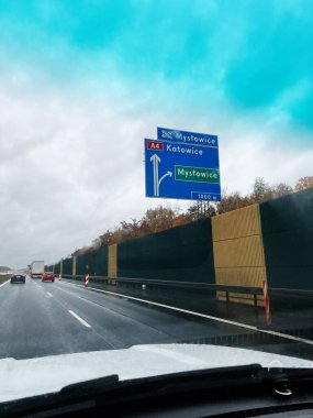 Arabalar ıslak bir Polonya otoyolunda ilerlerken, Mysowice Katowice ve Mystowice 'ı gösteren büyük mavi bir tabelayı geçerler..