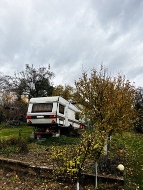 Sonbahar arka bahçesinde park etmiş bir karavan. Ağaçlar, yerde yapraklar ve gri bulutlu bir gökyüzü..