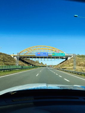 Üstteki kemer köprüsüne ve mavi yol işaretlerine doğru giden bir arabadan temiz bir otoyol manzarası..