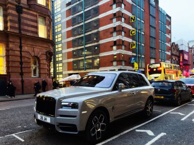 Manchester, İngiltere - 14 Ocak 2026: Kırmızı tuğlalı mimari ve parlak bir çift katlı otobüsün yanında kalabalık bir şehir caddesinde lüks bir Rolls-Royce Cullinan SUV dolaşır.