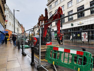 Manchester, İngiltere - 21 Ocak 2026: Manchester şehir merkezindeki kentsel inşaat sahnesi kırmızı Kazıcı vinç, eskrim ve geçen yayaları gösteriyor.