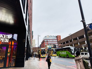 Manchester, İngiltere - 21 Ocak 2026: Manchester 'da yeşil Flixbus, sarı otobüsler, yayalar ve mağazaların yer aldığı işlek şehir caddesi.