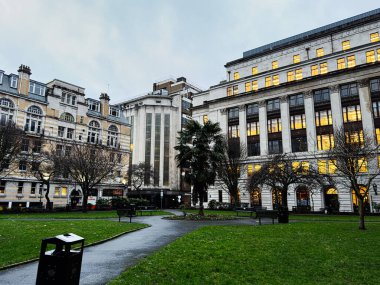 Manchester, İngiltere - 21 Ocak 2026: Büyük tarihi binalar ve alacakaranlıkta parlayan pencerelerle çerçevelenmiş sakin bir Manchester park alanı.