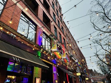 Manchester, İngiltere - 22 Ocak 2026: Festivalsel ışıklar, gökkuşağı LGBTQ bayrakları, tuğla perdeler ve açık hava mekanlarının yer aldığı hareketli Manchester sokak sahnesi.