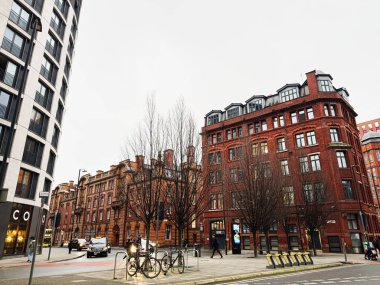Manchester, İngiltere - 22 Ocak 2026: Manchester 'da kırmızı tuğlalı iQ Hollingworth House cephelerinin, yapraksız ağaçların, bisikletlerin ve yayaların yer aldığı şehir sahnesi.