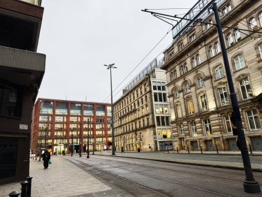 Manchester, İngiltere - 22 Ocak 2026: Manchester 'da tramvay rayları, tarihi cepheler ve modern kırmızı tuğlalı ofis bloğu ile sokak manzarası.
