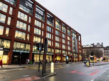 Manchester, İngiltere - 22 Ocak 2026: Tuğla ofis binası, cam pencereler, yayalar ve otobüslerin yer aldığı yoğun bir Manchester sokak sahnesi.