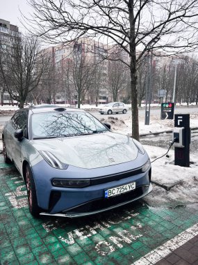Lviv, Ukrayna - 07 Şubat 2026: Gösterişli elektrikli Zeekr 001 sedan çıplak ağaçlar ve modern binalarla karlı bir şehir ortamında şarj oluyor.