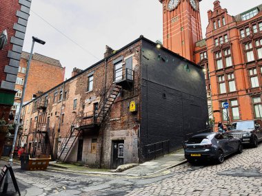 Manchester, İngiltere - 24 Ocak 2026: Kentsel Manchester sokağı tuğla binalar, yangın çıkışları, kaldırım taşı yollar ve arka planda saat kulesi.