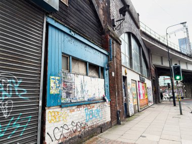 Manchester, İngiltere - 24 Ocak 2026: Manchester 'da dükkan vitrinleri ve yoğun kaldırım boyunca uzanan bir demiryolu üstgeçidi içeren cesur bir şehir caddesi.