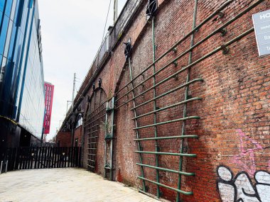 Manchester, İngiltere - 24 Ocak 2026: Manchester 'da yeşil parmaklıklar, graffitiler ve modern cam binalarla süslenmiş tuğla bir duvarın yanında dar bir sokak.
