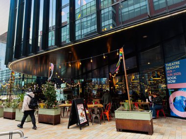 Manchester, İngiltere - 24 Ocak 2026: İnsanlar Manchester 'da cam cepheli bir binanın yanındaki sıcak sicim ışıkları altında ev yapımı sanat merkezinin keyfini çıkarıyorlar.