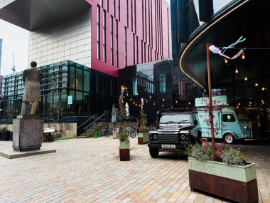 Manchester, İngiltere - 24 Ocak 2026: Manchester 'da Friedrich Engels heykeli, cam mimarisi ve ışık altında dekore edilmiş dağıtım kamyonlarının yer aldığı şehir sahnesi.